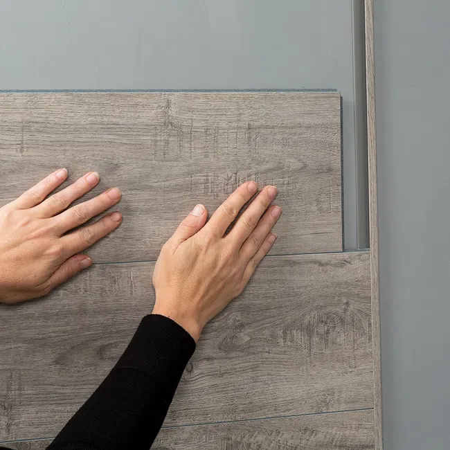 Hands installing gray wood laminate flooring on a smooth gray surface in a close-up view.