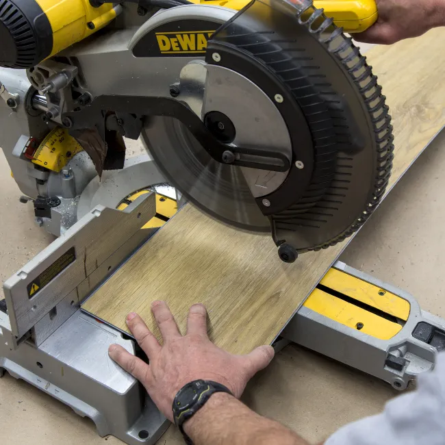 Person using a DeWalt miter saw to cut a wooden plank on a workshop table with safety precautions.