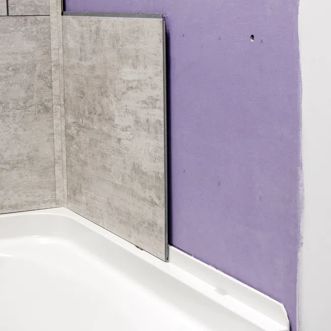 Partially installed grey wall tiles above a white bathtub next to an unfinished purple drywall corner.