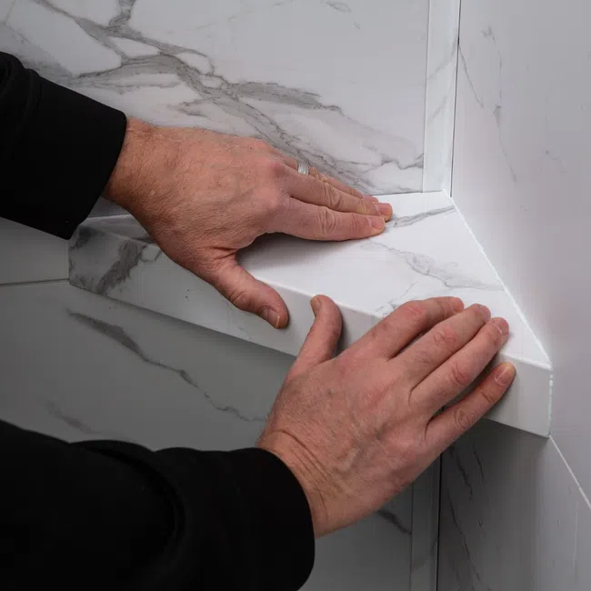 Hands installing a white marble-patterned shelf or ledge in a corner against matching wall tiles.
