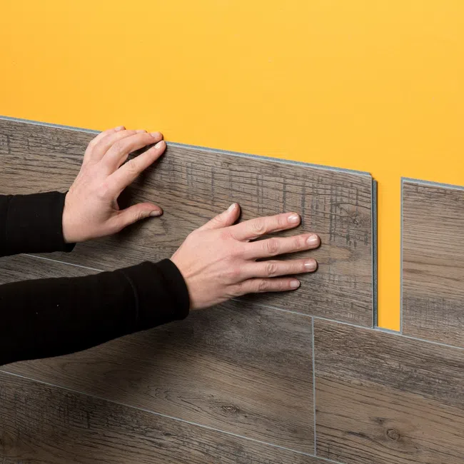 Hands installing wood-look vinyl planks on a yellow wall during interior renovation work.