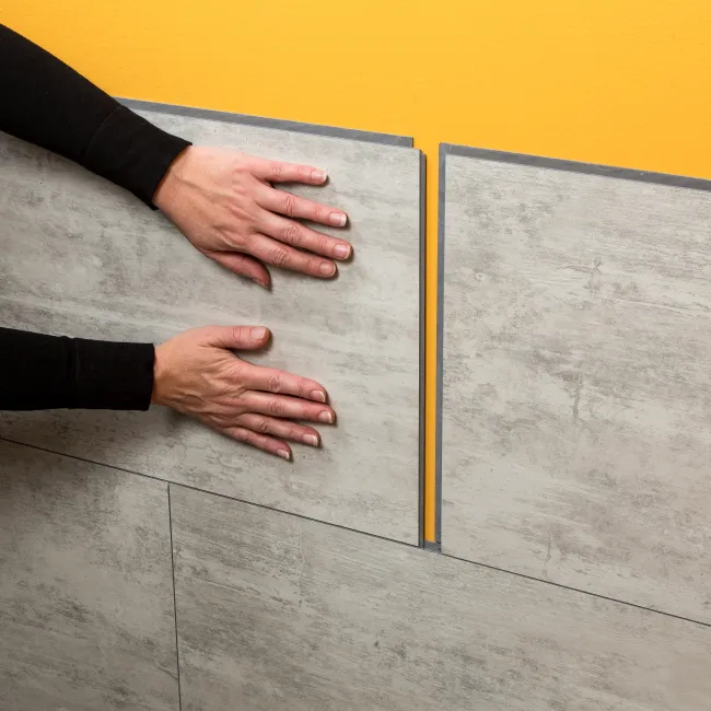 Person installing large gray ceramic tiles on a yellow wall, hands pressing the tile into place.