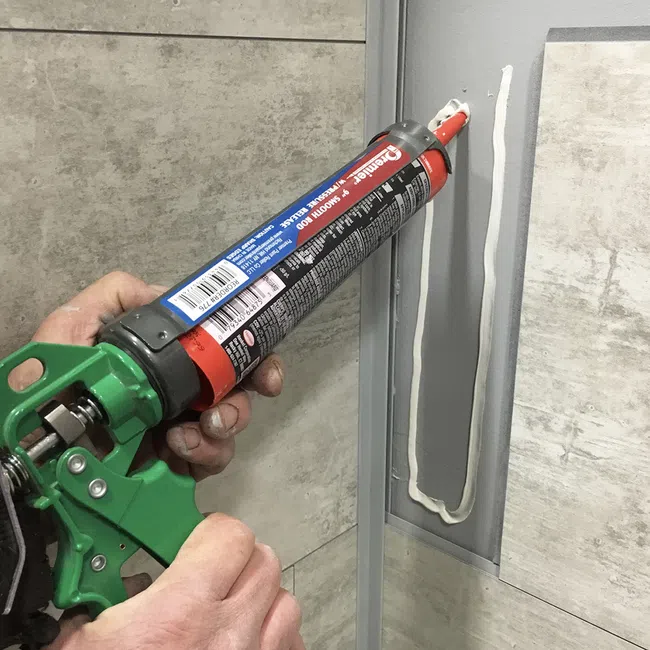 Hand applying white sealant with a green caulking gun between tiles in a bathroom corner.