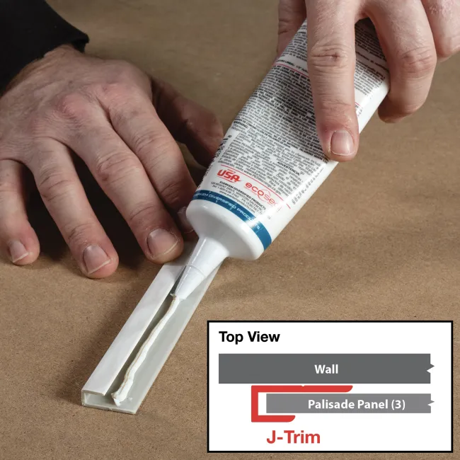 Hand applying white adhesive sealant from a tube onto a white J-Trim molding piece on a brown surface.