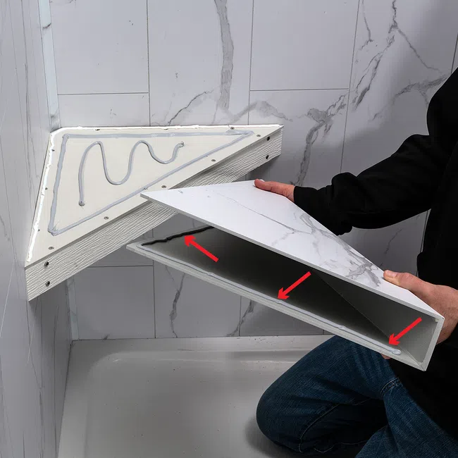 Man installing a triangular marble-patterned shelf with adhesive in a bathroom corner with tiled walls.