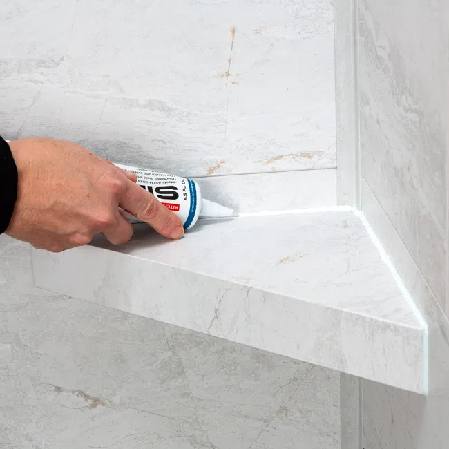 Hand applying white silicone sealant to a corner joint of a marble shelf and wall in a bathroom.