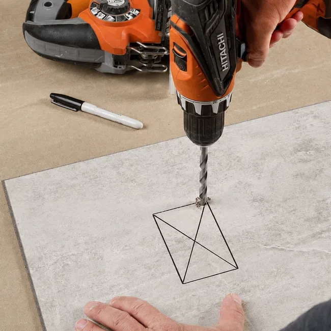 Person using an orange Hitachi power drill to drill a marked tile on a workbench with tools nearby.