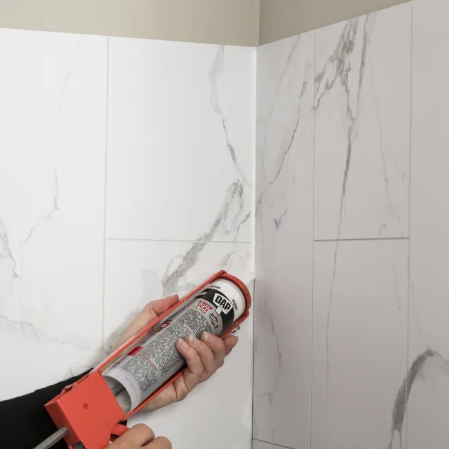 Person applying sealant to the corner of white marble-effect tiled walls using a caulking gun.