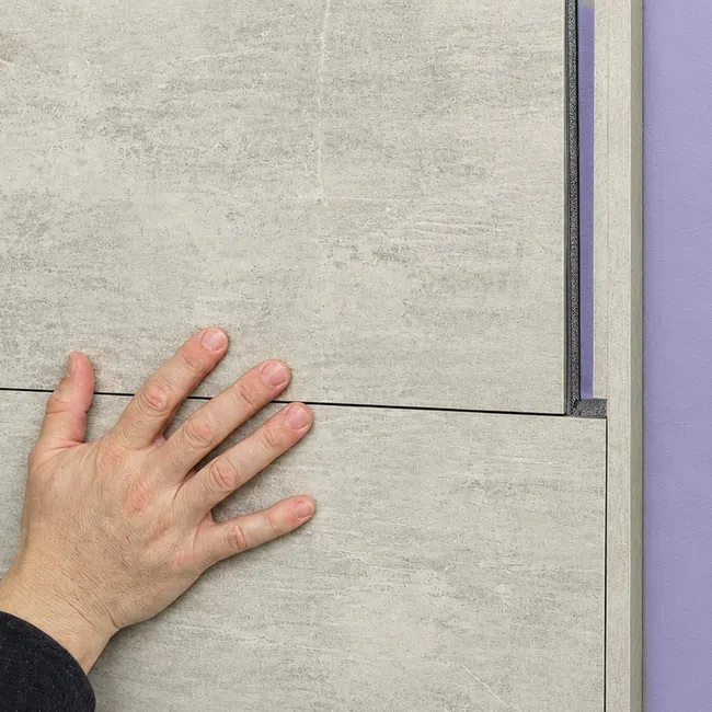 Hand installing large gray wall tile panels against a purple background, showing smooth texture and precise fitting.