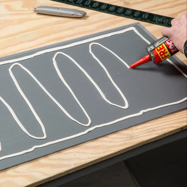 Applying white construction adhesive in zigzag pattern on gray panel on wooden workbench with tools nearby