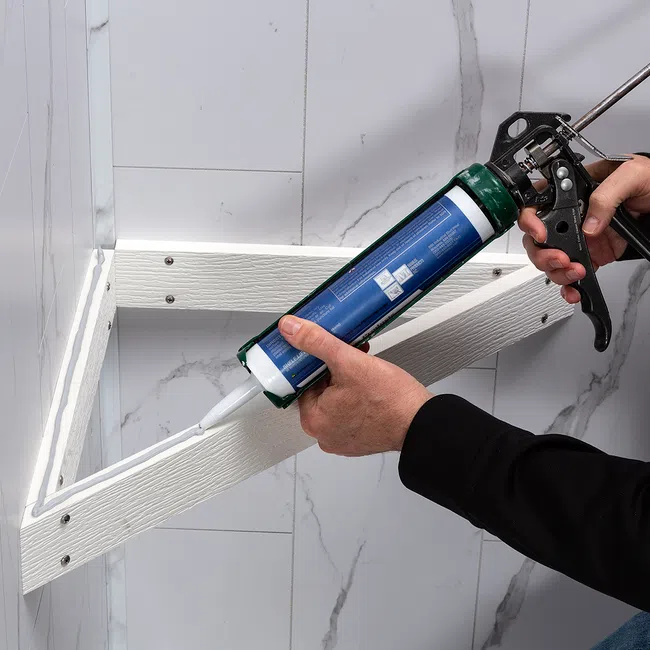 Person applying white sealant with a caulking gun to a corner joint on a tiled wall around wooden trim.