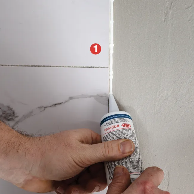 Hands applying white sealant to the gap between marble tiles and a wall corner for waterproofing.