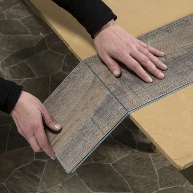 Person fitting gray wood-look vinyl flooring planks on a table over stone-tiled floor background