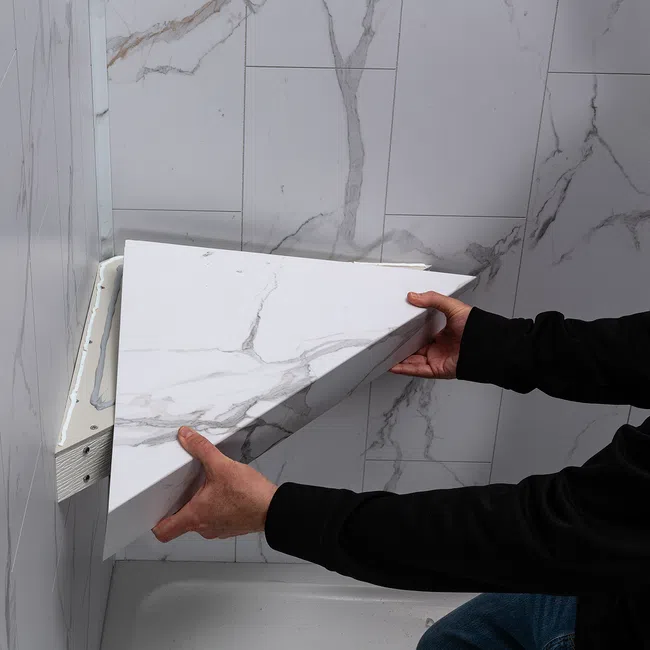 Person installing a white marble-patterned triangular shelf in a corner with marble wall tiles.