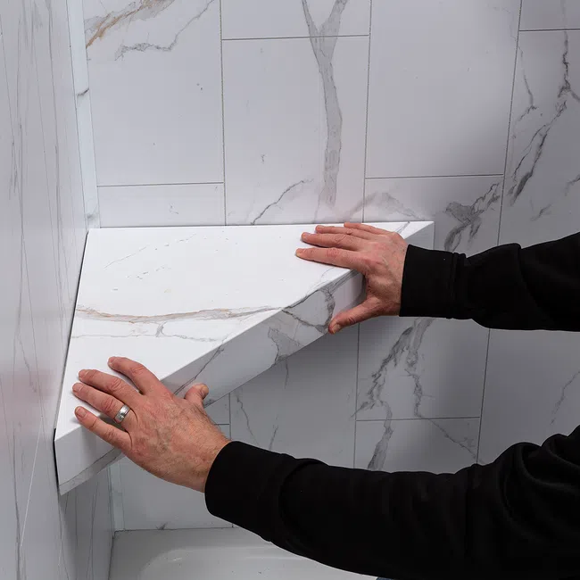 Hands installing a white marble-patterned triangular shelf on a matching tiled wall in a bathroom corner.