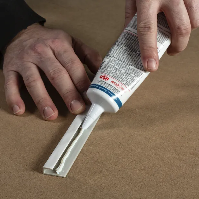Person applying white adhesive from a tube onto a hinge on a brown surface.
