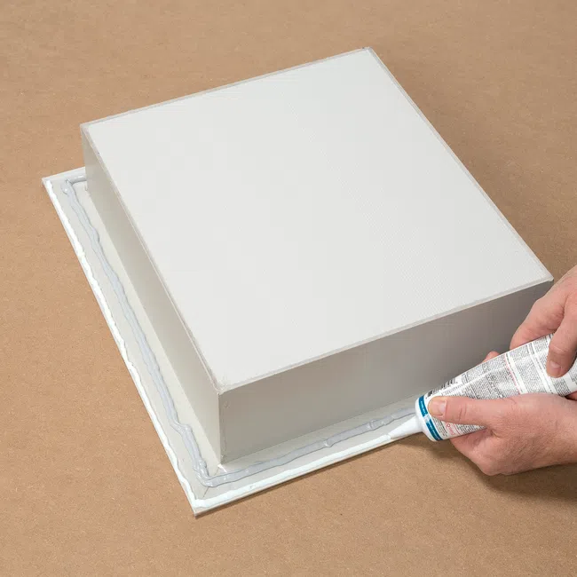 Hands applying adhesive sealant around the base of a white rectangular box on a brown surface.