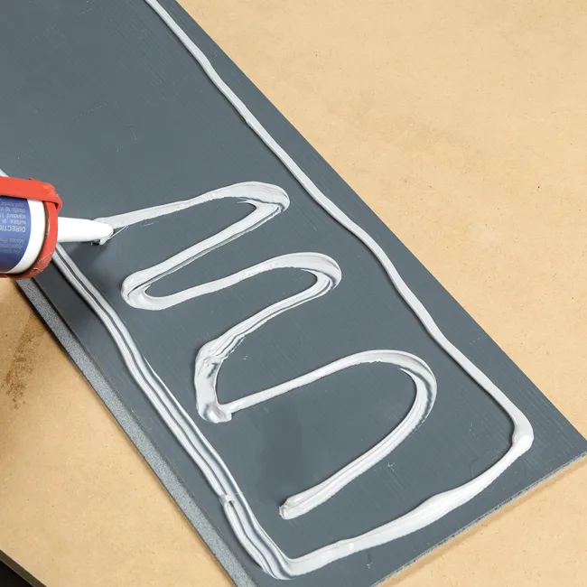 Close-up of white adhesive applied in a zigzag pattern on a dark gray wooden plank on a beige surface.