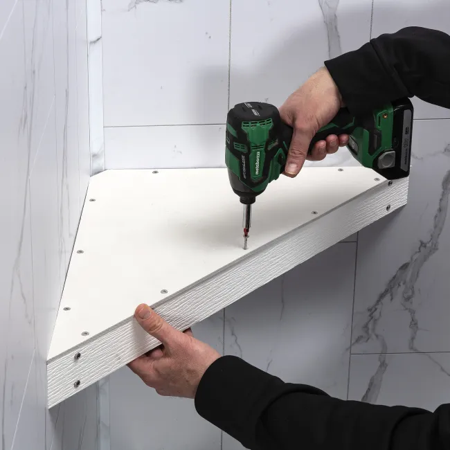Person using a green cordless drill to fasten a white triangular shelf against a marble wall.