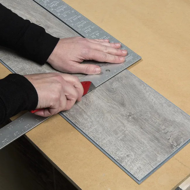 Person cutting vinyl plank flooring with a utility knife using a metal ruler for precision on a work table.
