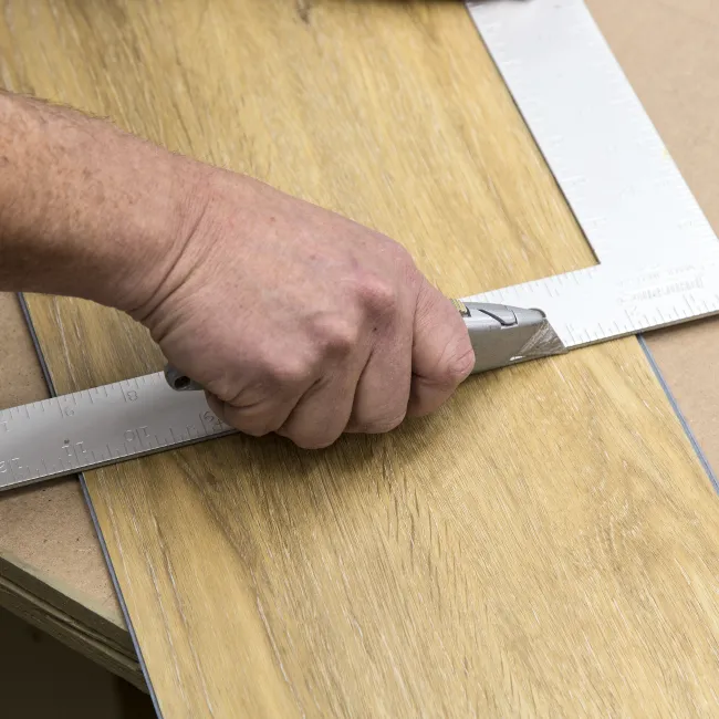 Hand cutting wood laminate flooring with a utility knife using a metal square ruler for precision.