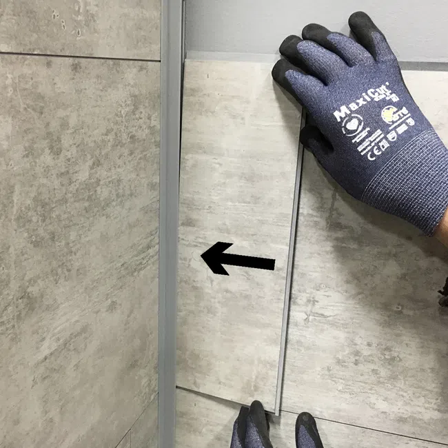 Hand wearing protective gloves installing or aligning rectangular stone tile with black arrow on wall