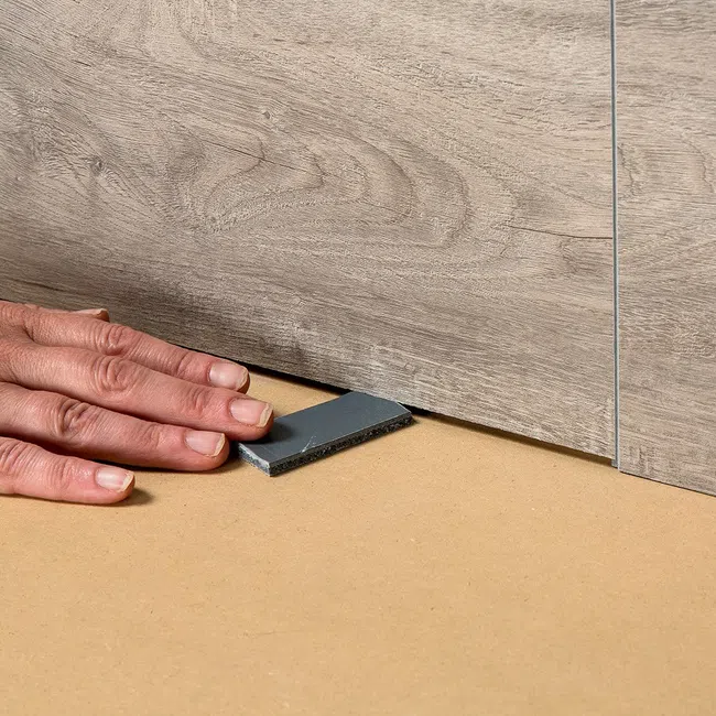 Hand placing a leveling spacer under wood grain floor panels during flooring installation for alignment.