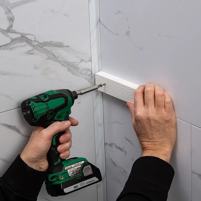Person using a green cordless drill to screw a white wooden board into a marble wall corner.