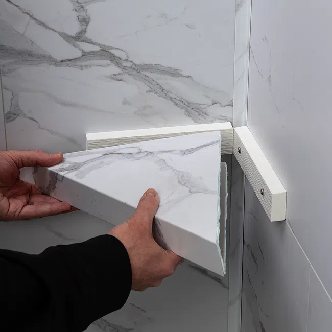 Hands installing a triangular marble-patterned shelf corner on a wall with marble tiles and white support brackets.
