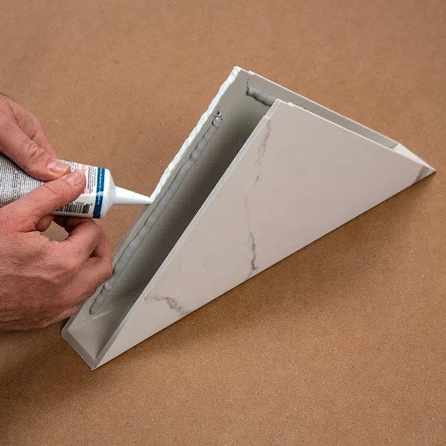 Person applying adhesive glue to the edge of a triangular marble-patterned object on a brown surface.