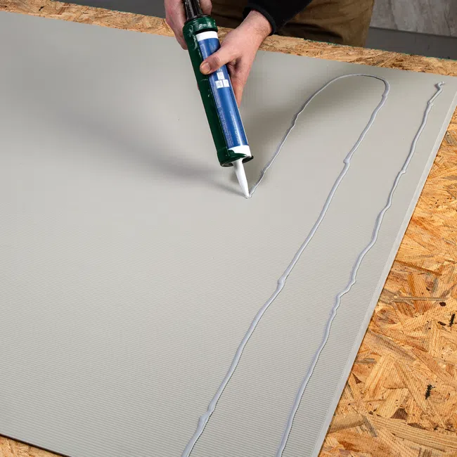 Person applying adhesive with caulking gun onto a large gray panel on a wooden surface during construction work.