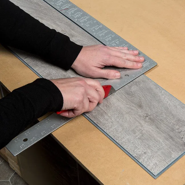 Person cutting vinyl plank flooring with utility knife and metal ruler on wooden surface
