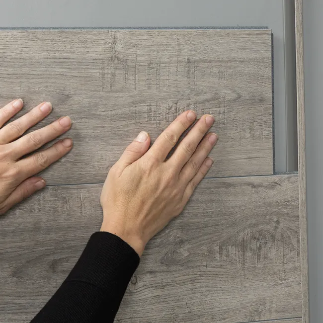 Hands installing gray wood-look laminate flooring planks over a gray subfloor surface indoors