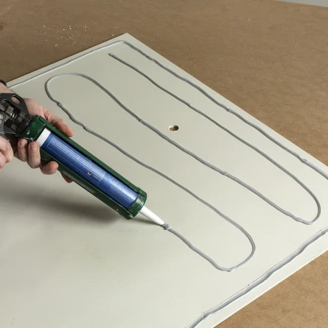 Person applying gray adhesive with caulking gun on white panel with a drilled hole on brown surface.