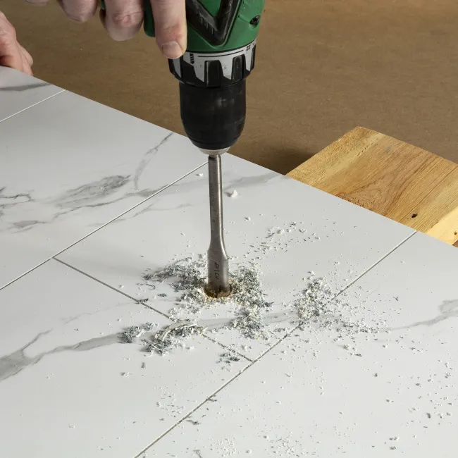 Handheld electric drill making a hole in white marble tile with dust and debris scattered around.
