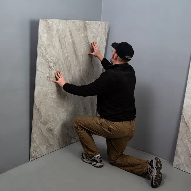 Man kneeling to install large gray marble tile against a plain gray corner wall indoors.