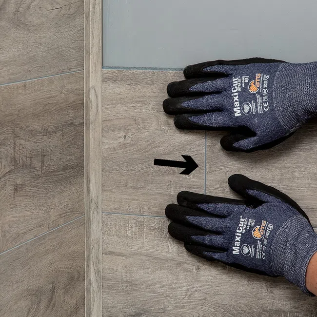 Hands wearing MaxiCut safety gloves placing wood-look floor tiles with alignment arrow on surface.