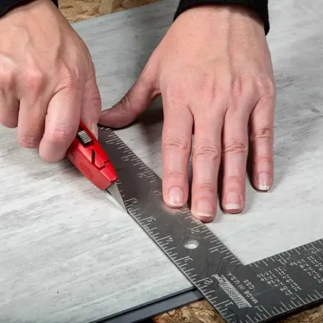 Hands using a red utility knife and metal square ruler to cut a gray tile precisely on a work surface.