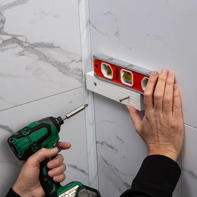 Person using a green cordless drill to screw a white wooden board to a tiled wall, checking level with a red spirit level.