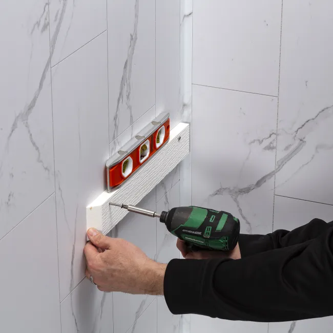Man using a power drill to screw a wooden plank on a white tiled wall with a level tool on top