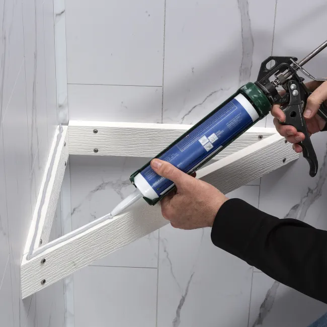 Person applying white silicone sealant with caulking gun on white wooden frame in tiled corner.