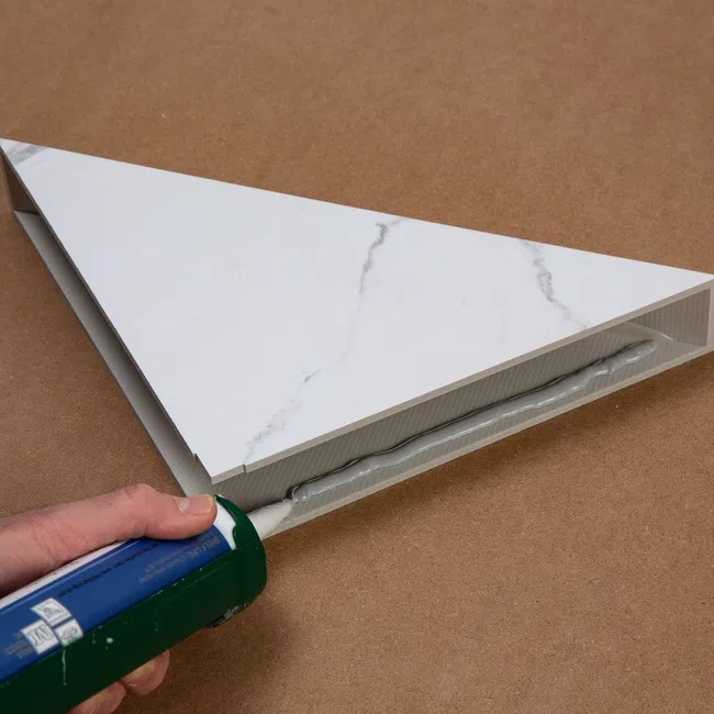 Hand applying adhesive with a caulking gun to the underside of a triangular marble-patterned tile on a brown surface.