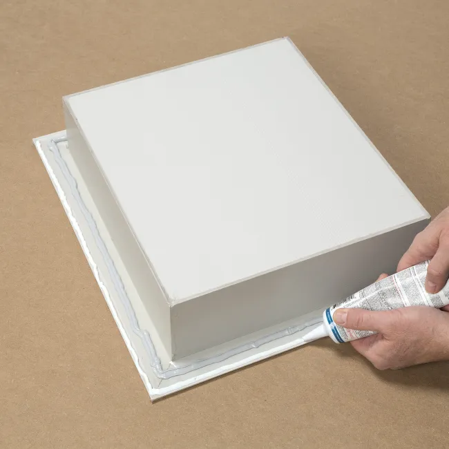 Person applying white adhesive sealant around the base of a white rectangular object on a brown surface.