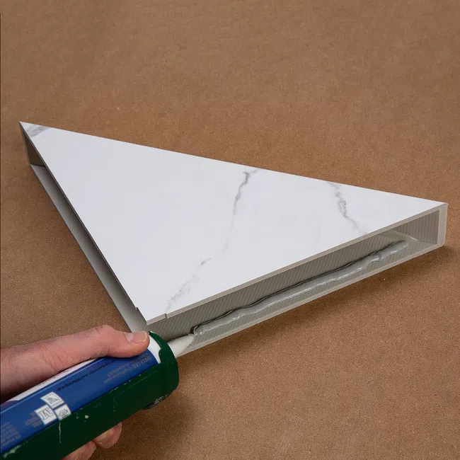 Hand applying adhesive with caulking gun to secure white marble-patterned triangular panel on brown surface.