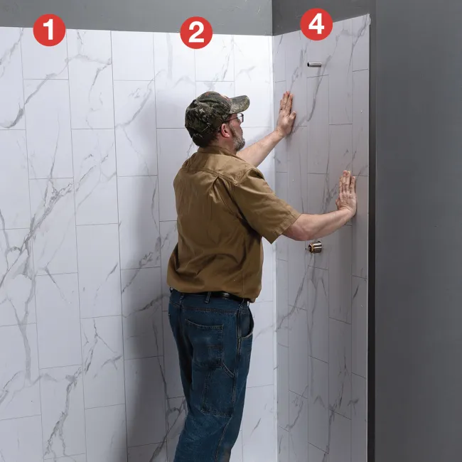 Man installing large white marble-effect wall panels in a corner bathroom space with gray walls.