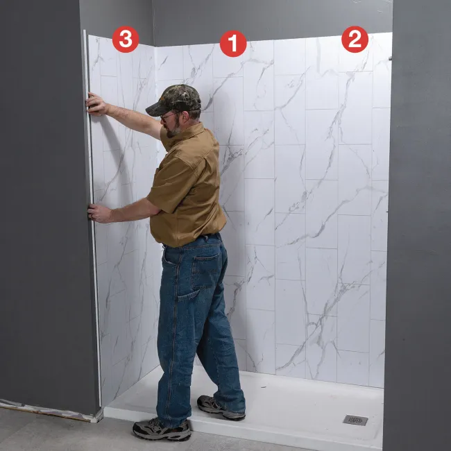 Man installing white marble-patterned shower wall panels in a corner bathroom space with numbered steps