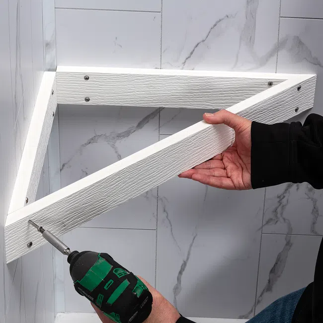 Person using a cordless drill to install a white triangular wooden shelf on a marble tiled wall.