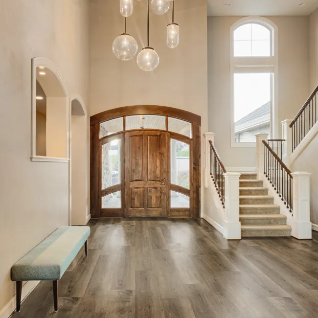 Spacious entryway with wooden front door, modern pendant lights, carpeted stairs, and a blue bench on wood flooring