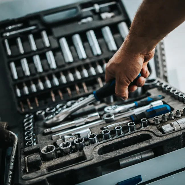 Hand holding a ratchet wrench over an organized socket set in a toolbox drawer with various tools.
