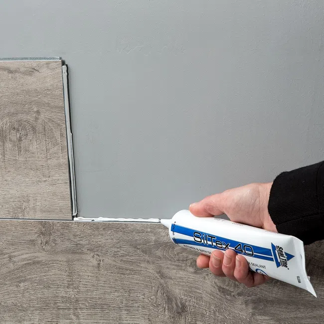 Person applying white sealant from a tube to gap between wood-patterned floor tiles and a gray wall.